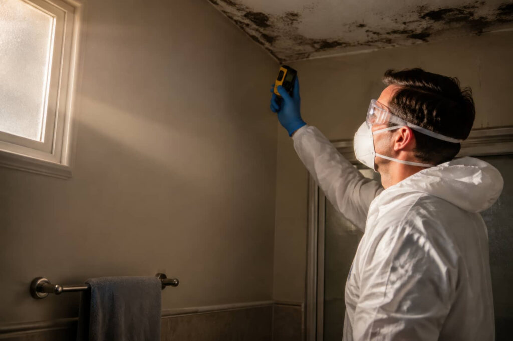 Person in protective gear inspecting black mold on a bathroom ceiling.