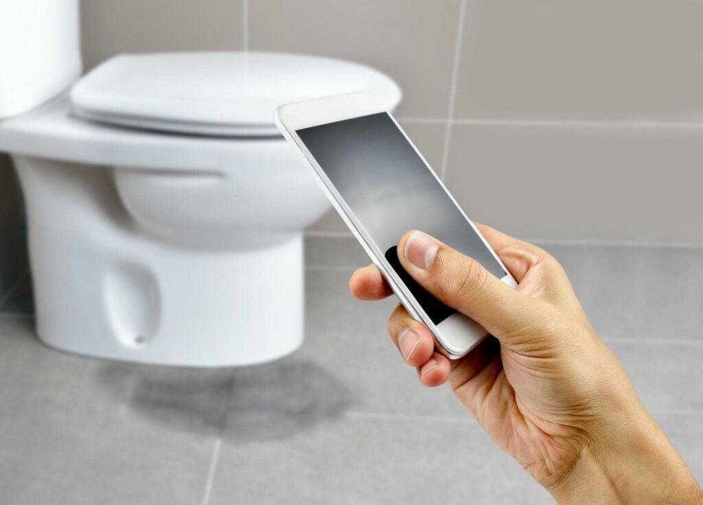 Hand holding a smartphone in front of a closed toilet in a bathroom.